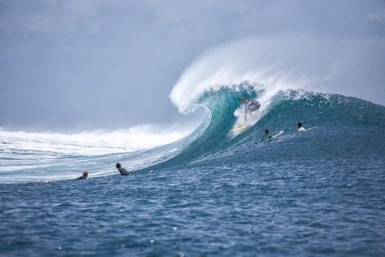 Surfeur lancé dans une immense vague bleue, sous le regard d’autres surfers dans l’eau, symbolisant une pratique intensive et engagée du sport aquatique.