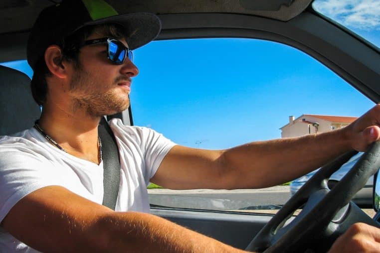 Jeune homme au volant d’une voiture, ceinture attachée, portant des lunettes de soleil sombres en conduisant sous un ciel bleu lumineux.