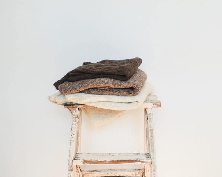 Pile de pulls en maille beige, marron et gris soigneusement pliés sur un escabeau en bois patiné devant un mur blanc.