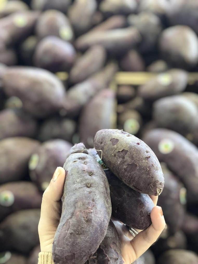 Patates douces violettes entières disposées dans des cagettes sur un étal de marché, montrant leur peau foncée et leur forme allongée.