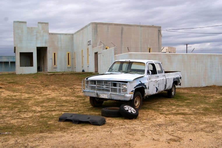 Vehicule militaire d’entraînement abandonné devant un bâtiment gris sur un terrain sec, symbolisant une zone d’exercices.