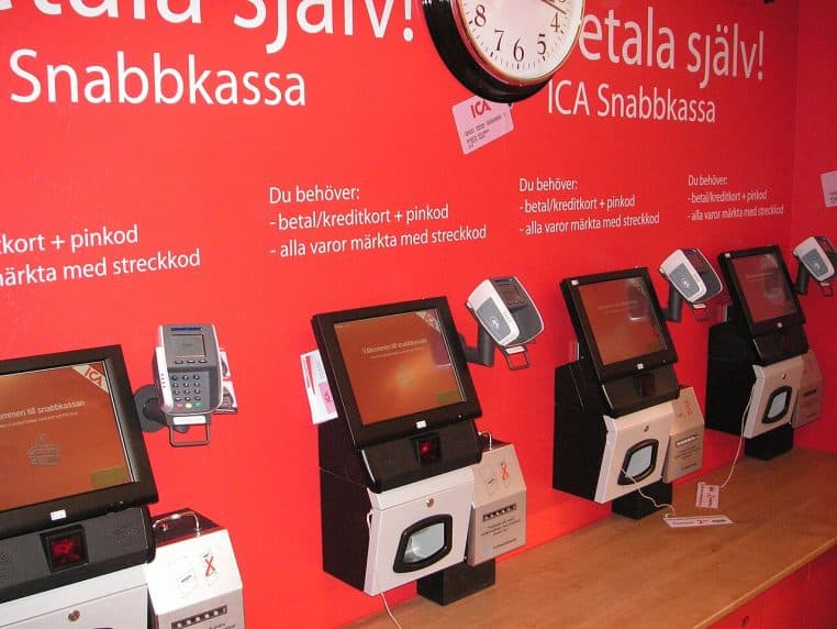 Caisse de libre-service dans un supermarché, avec écran, scanner et zone de dépôt des articles visibles au premier plan.