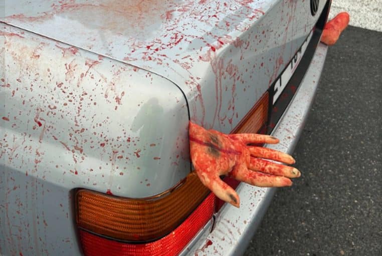 Voiture grise éclaboussée de faux sang, mains factices coincées dans le coffre, gros plan sur le feu arrière gauche.