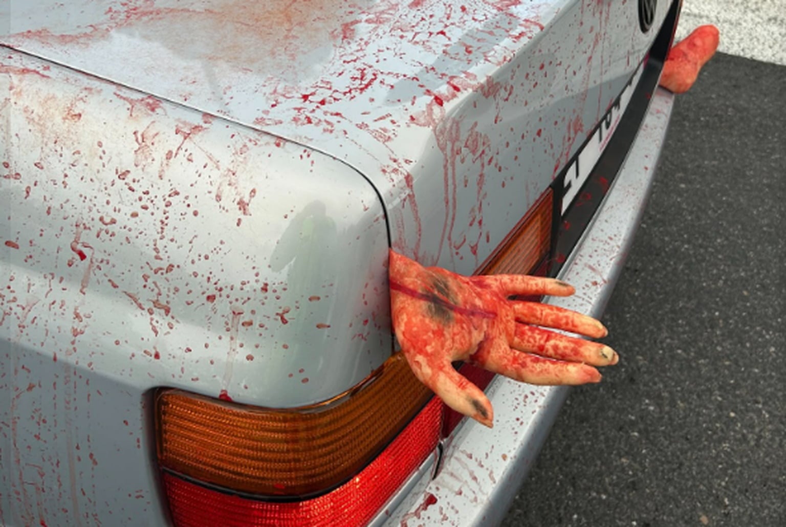 Voiture grise éclaboussée de faux sang, mains factices coincées dans le coffre, gros plan sur le feu arrière gauche.