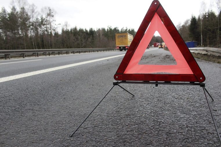 Triangle de sécurité orange installé sur la bande d’arrêt d’urgence d’une autoroute, avec plusieurs poids lourds visibles à l’horizon