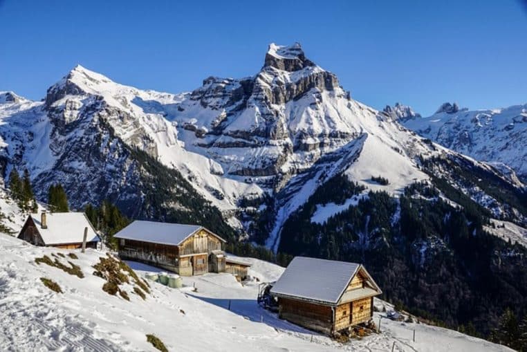 Chalets de montagne enneigés face à un sommet escarpé des Alpes, baignés par un soleil d’hiver dans une atmosphère très claire.