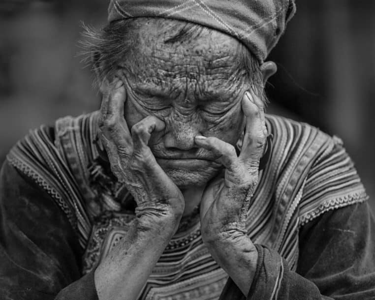 Portrait en noir et blanc d’une femme âgée assise, yeux fermés, visage marqué, tenant sa tête entre ses mains fatiguées.