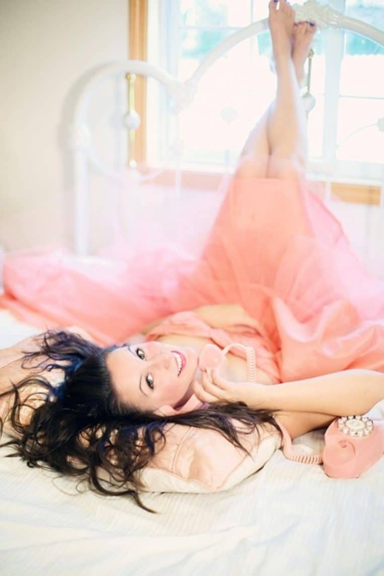Femme allongée sur un lit, robe rose et téléphone vintage à la main, souriant en regardant vers l’objectif, ambiance détente.