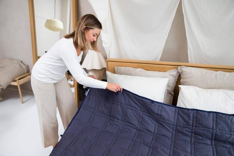 Femme en tenue d’intérieur étendant soigneusement une couverture épaisse bleu nuit sur un lit double dans une chambre lumineuse et épurée.