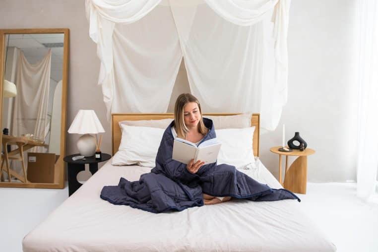 Femme lisant paisiblement assise sur un lit double, enveloppée dans une couverture alourdie bleu nuit dans une chambre claire au décor minimaliste.