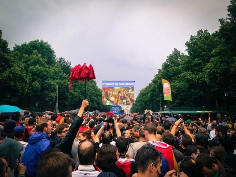 Supporters rassemblés en nombre dans une fan zone en plein air devant un écran géant diffusant un match de Coupe du monde, banderoles et drapeaux brandis au-dessus des têtes.