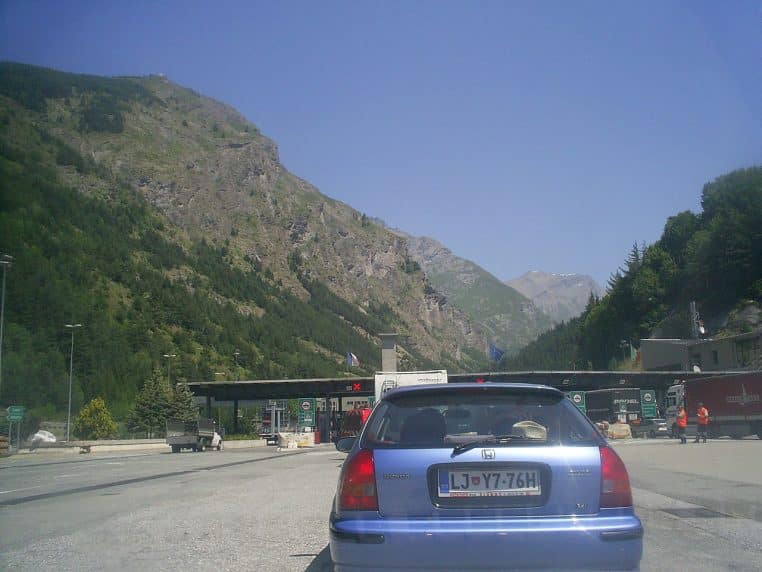 File de voitures devant un péage sur l’A32 côté Italie, avec montagnes en arrière-plan et ciel dégagé.