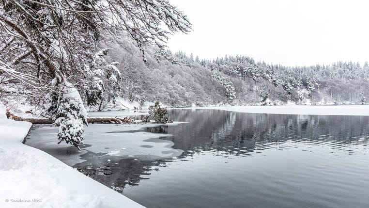 Paysage d’hiver au lac Pavin dans le Massif central, avec berge enneigée, eau sombre et arbres givrés sous un ciel froid et lumineux.