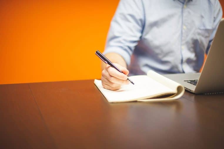 Homme prenant des notes devant un ordinateur portable sur une table, scène de concentration et d’engagement au travail.