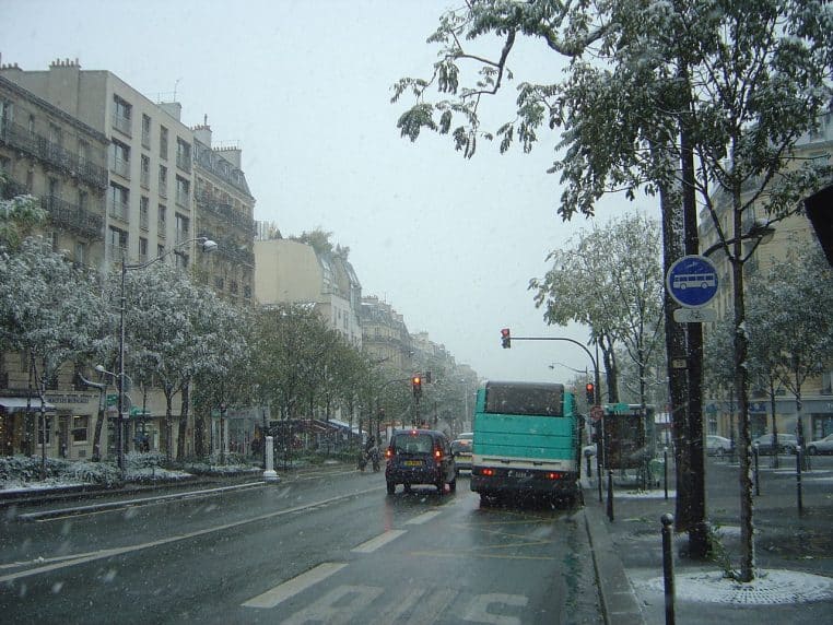 Avenue parisienne sous une chute de neige, circulation et bus au premier plan, arbres et immeubles blanchis en arrière-plan.