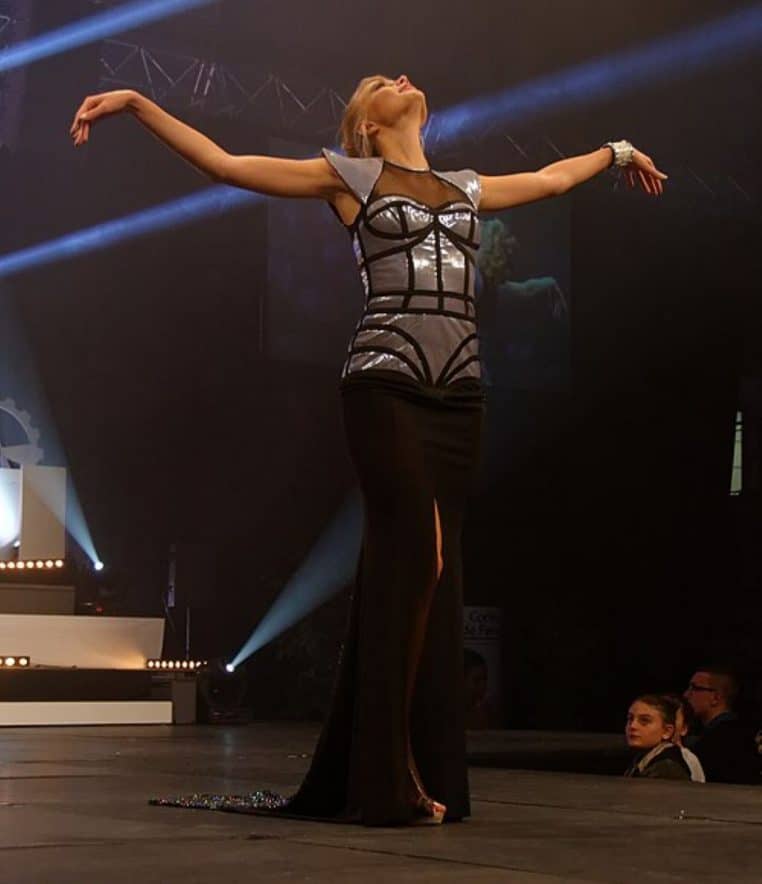 Une Miss en robe longue noire au bustier métallisé pose bras écartés au centre de la scène, éclairée par les projecteurs d’un défilé de concours de beauté.