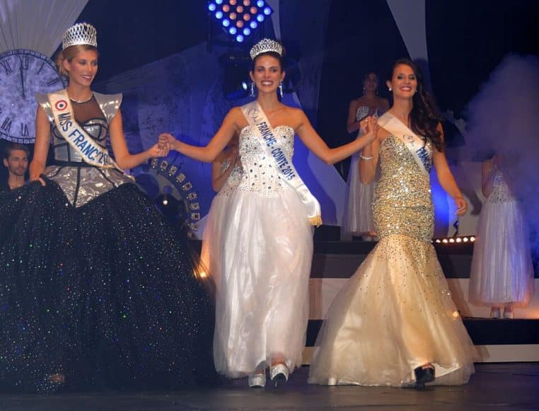 Sur la scène illuminée d’un concours régional, Miss France 2015 et les lauréates défilent main dans la main en robes de bal sous les jeux de lumière.