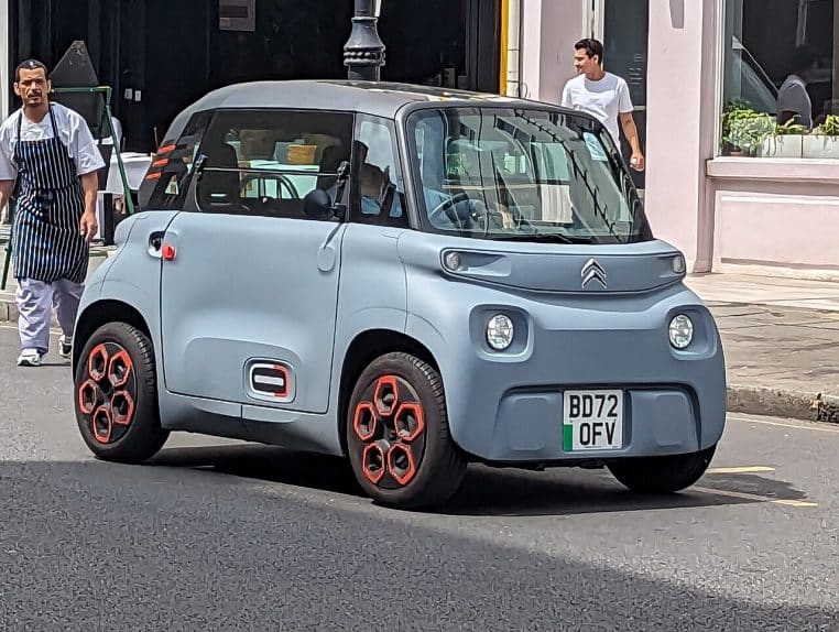 Citroën Ami électrique gris-bleu circulant en ville, photographiée de profil dans une rue commerçante animée, avec des passants sur le trottoir en arrière-plan.