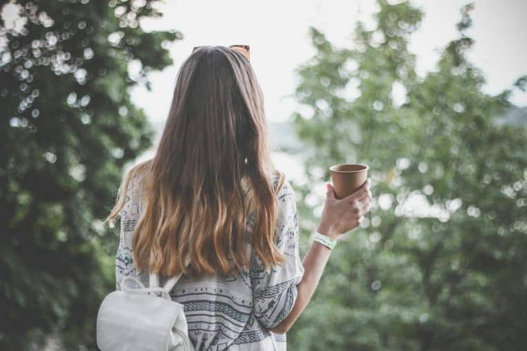 Back view of a young woman with long hair holding a coffee cup outdoors in a tranquil park.