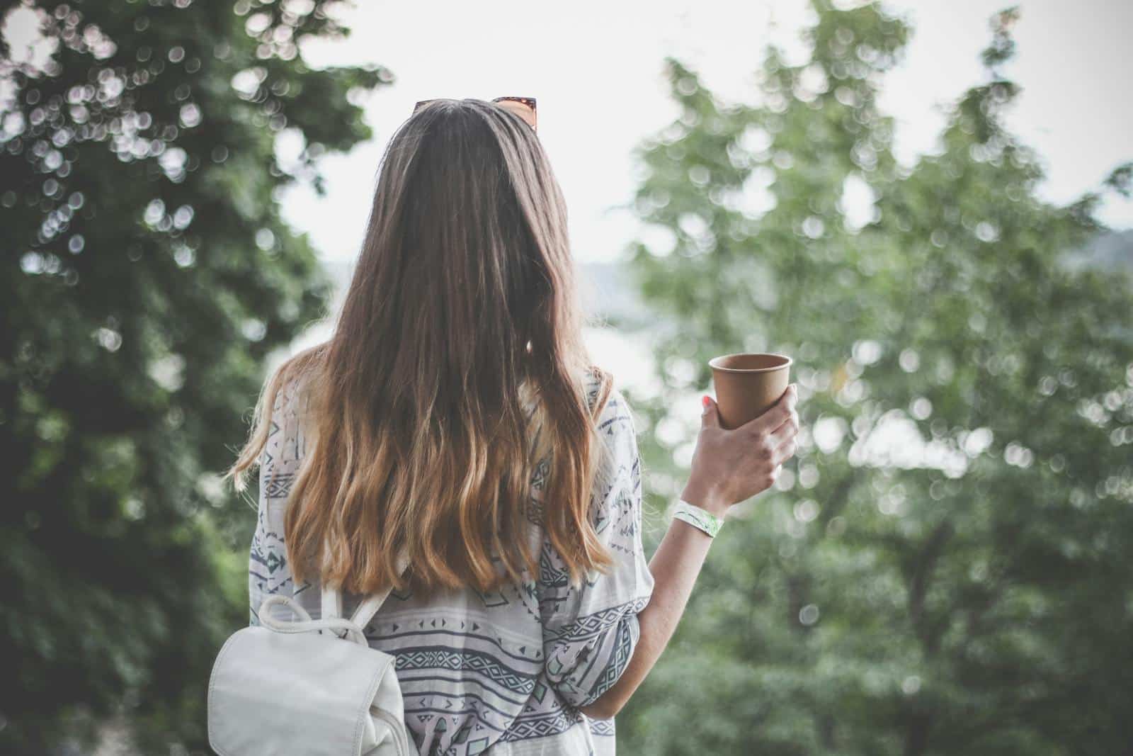 Back view of a young woman with long hair holding a coffee cup outdoors in a tranquil park.