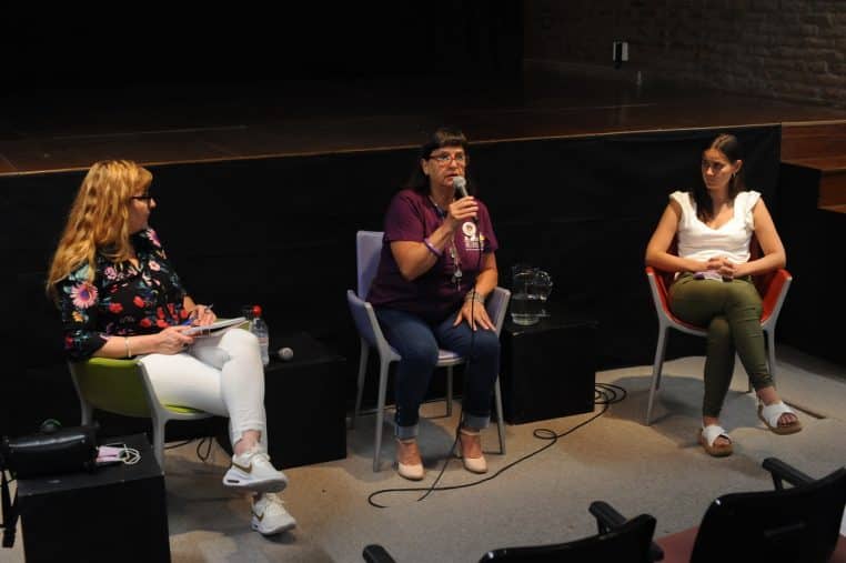 Trois femmes assises sur scène discutent lors d’un débat public, l’une tenant un micro pour répondre aux questions.