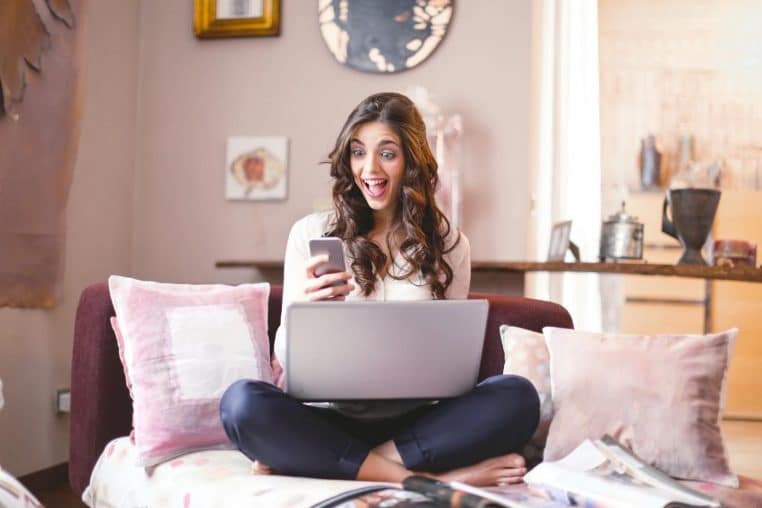 Smiling woman at home, using smartphone and laptop, expressing surprise and happiness.