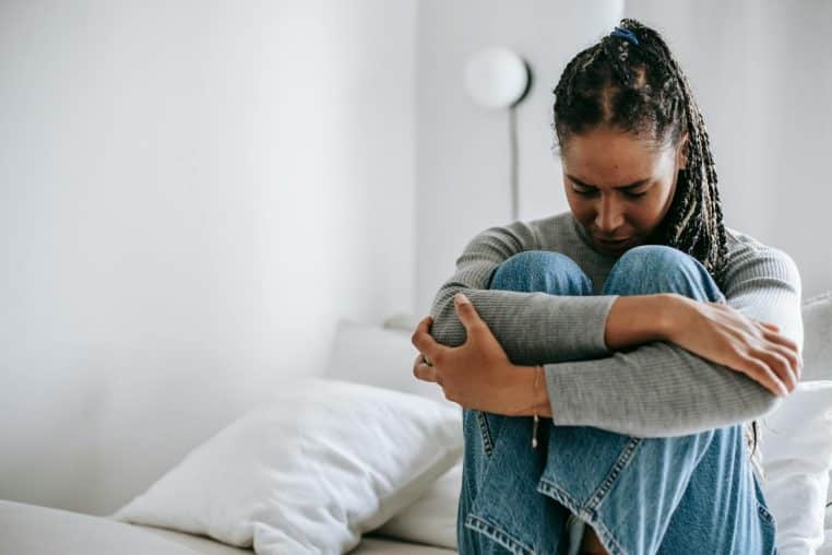 A young woman embraces her knees while sitting on a bed, appearing thoughtful and introspective.