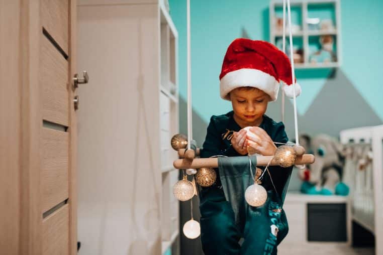 A child in a Santa hat sitting on a swing indoors, embracing Christmas decor.