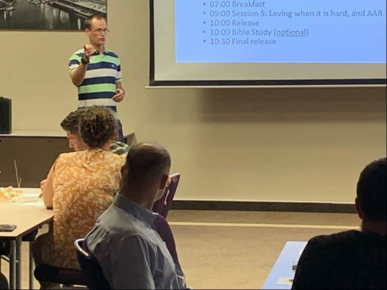 Un intervenant présente l’agenda d’une retraite de couples devant un écran, pendant qu’un groupe de participants l’écoute attentivement.