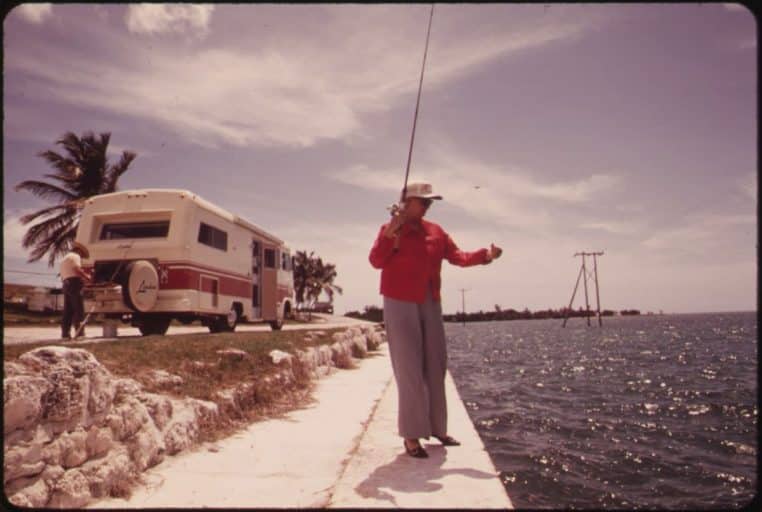 Couple de retraités près d’un camping-car, l’un pêchant au bord de la mer, l’autre s’affairant, symbole de liberté grâce à la pension.