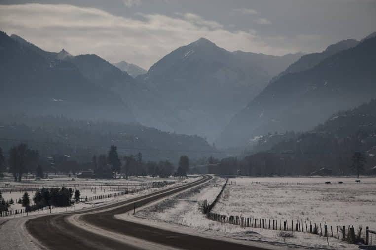 Route sinueuse bordée d’arbres enneigés menant vers des montagnes brumeuses sous un ciel d’hiver clair.