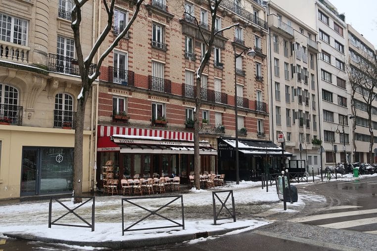 Avenue parisienne enneigée avec terrasses vides, façades d’immeubles et chaussée humide en fin d’après-midi.