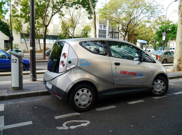 Petite voiture électrique grise Bolloré Bluecar branchée à une borne Autolib’ sur rue à Paris, entourée d’autres véhicules en stationnement.