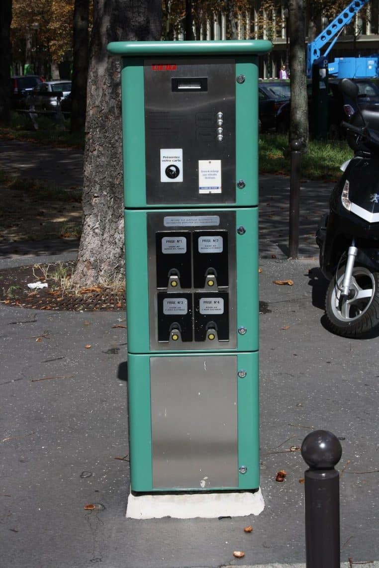 Borne de recharge pour véhicule électrique installée sur un trottoir parisien, avec voiture branchée au câble de charge en journée en ville.