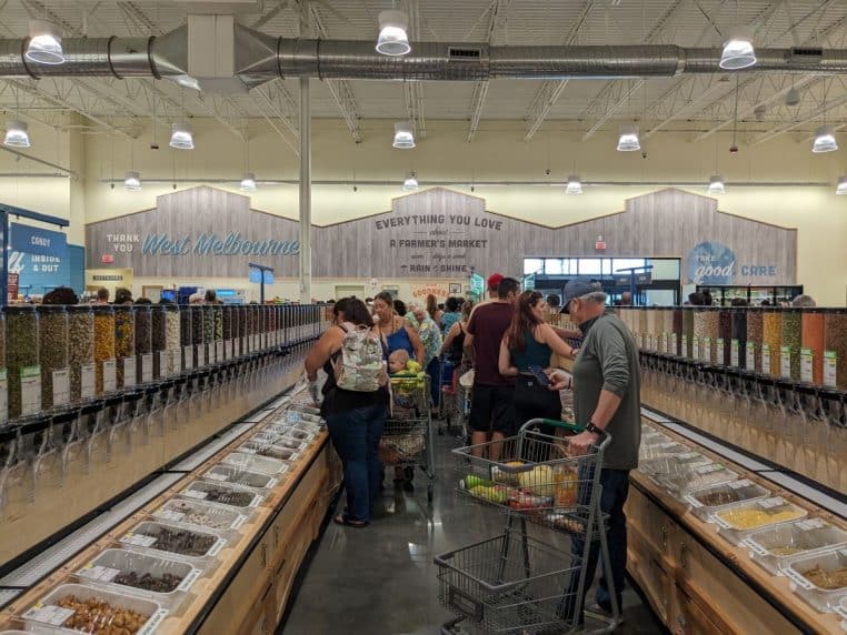 Allée de magasin avec distributeurs de vrac des deux côtés et clients qui remplissent leurs contenants et sacs