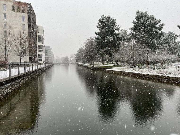 Canal du Parc Bühler à Bordeaux sous la neige, arbres givrés et immeubles modernes reflétés dans une eau sombre parsemée de flocons.