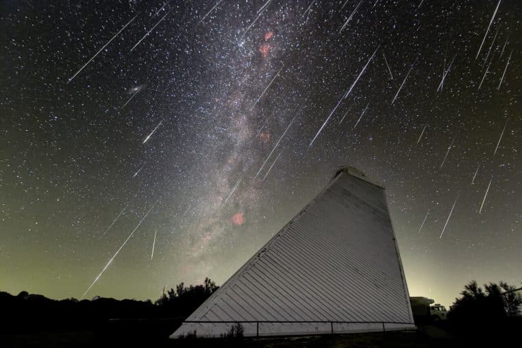 Large panorama de la Voie lactée avec des traînées lumineuses fines rappelant une pluie de météores, au-dessus d’un relief de collines sombres.