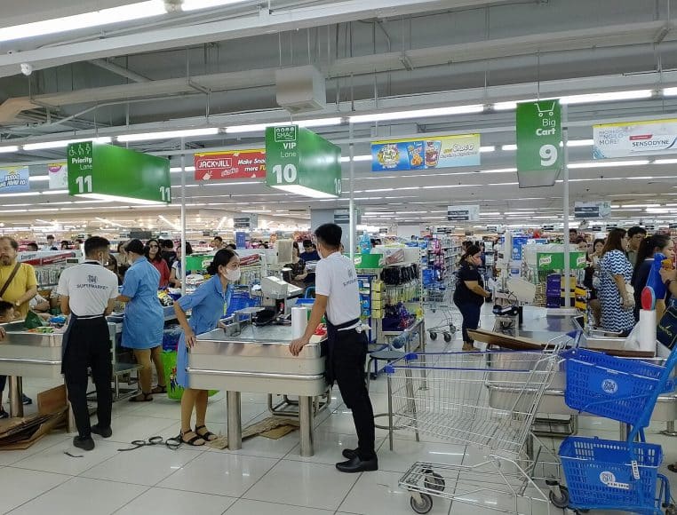 Hôtesses et hôtes de caisse travaillant dans une grande allée de supermarché bondée, avec plusieurs tapis en fonctionnement et des chariots alignés.