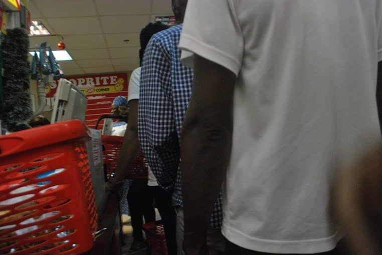 File de clients serrés avec paniers rouges dans un supermarché nigérian, attendant devant la caisse entre rayons et affiches colorées.