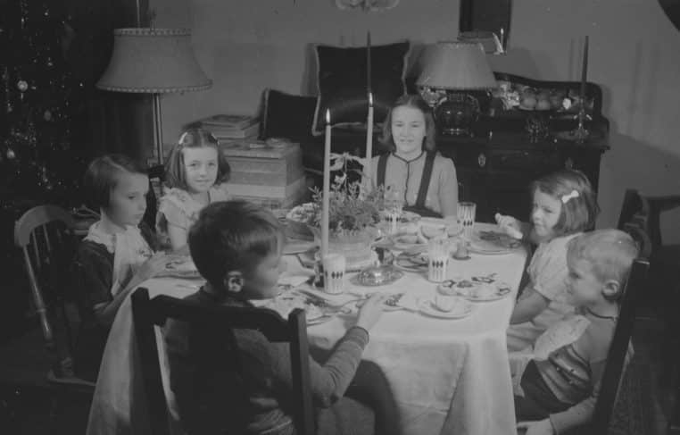 Groupe d’enfants assis autour d’une table de Noël en 1938, sourires timides et bougies au centre du repas festif.
