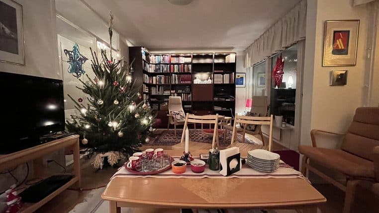 Salon scandinave en Suède avec sapin de Noël illuminé, table basse, fauteuils cosy et boissons chaudes posées à proximité.