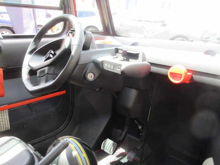 Vue de l’habitacle d’une Citroën Ami montrant le volant, le tableau de bord dépouillé et le support pour smartphone, avec des touches de couleur orange vives.