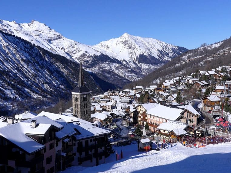 Vue sur le village enneigé de Saint-Martin-de-Belleville avec clocher, toits blancs et montagnes de Savoie sous un ciel bleu intense.