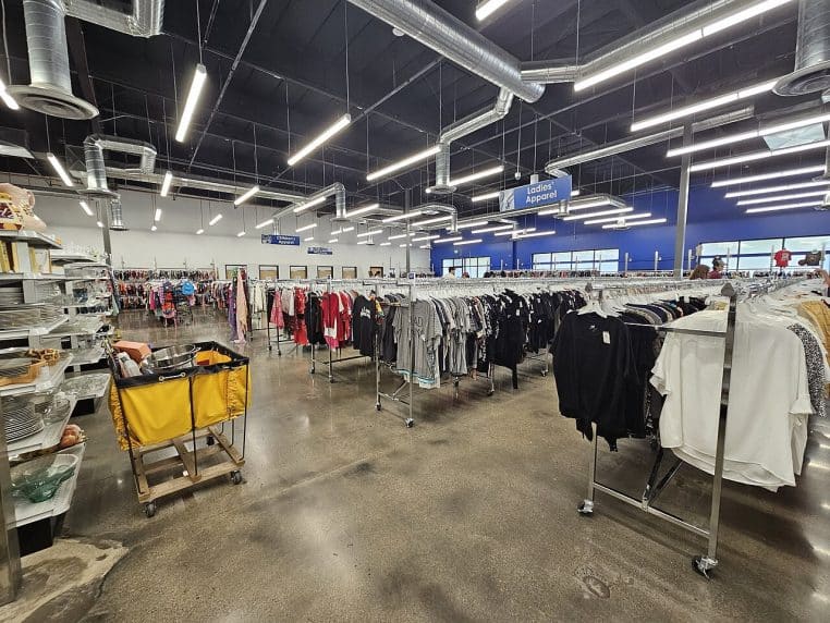 Intérieur d’une boutique de seconde main avec portants de vêtements, ambiance “chiner” et allées lumineuses.