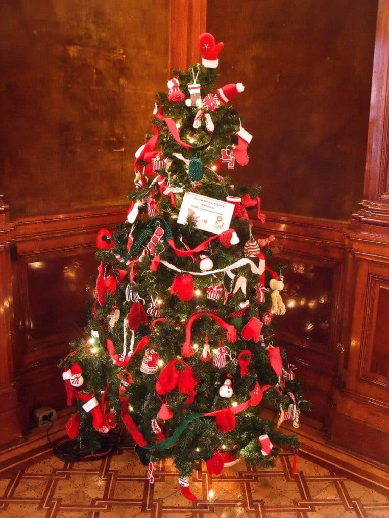 Grand sapin de Noël dressé dans un salon historique avec boiseries, meubles anciens et lumière naturelle traversant les fenêtres.
