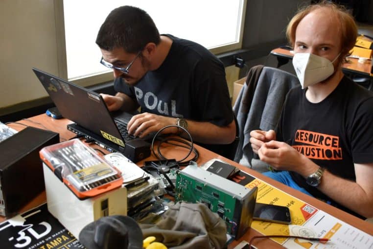 Deux personnes travaillent sur un ordinateur portable entouré de disques et de kits, ambiance atelier informatique.
