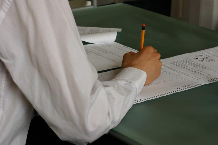 Élève en chemise blanche remplissant une grille de réponses sur une table d’examen, crayon à la main, dans une salle de classe silencieuse pendant une épreuve écrite du bac.