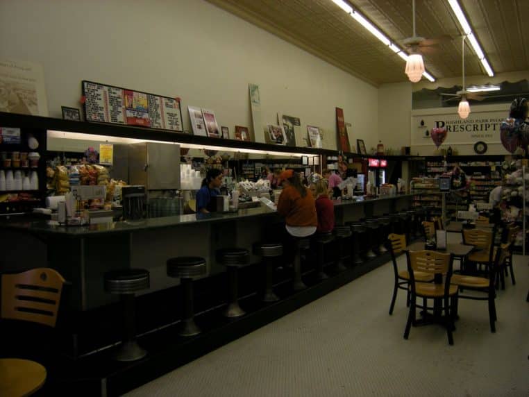 Intérieur d’une pharmacie avec long comptoir, tabourets et clients, éclairage de plafond et rayonnages