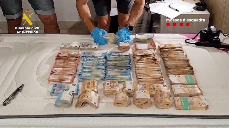 Agents en gants bleus manipulant des piles de billets en euros alignées sur un lit, illustrant l’argent liquide saisi lors d’une opération des autorités.