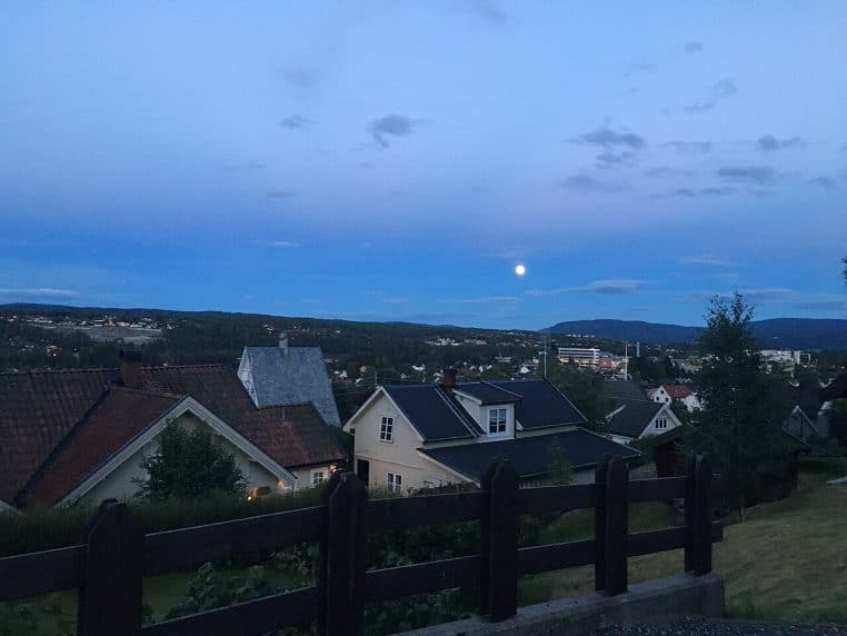 Panorama d’un quartier de maisons avec toits en pente au crépuscule, une pleine Lune blanche se détachant dans le ciel bleu sur fond de collines.